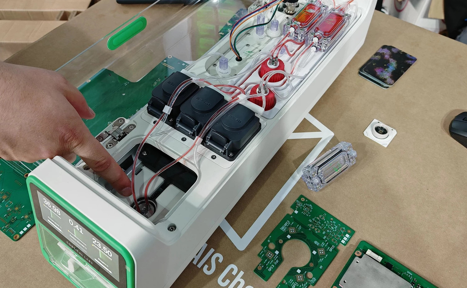Interior de um biocomputador com bombas peristálticas, tubos e reservatórios; mão aponta para o poço de acesso enquanto placas de circuito estão sobre a bancada.