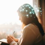 Mulher jovem escrevendo em diário pessoal perto da janela, luz natural da manhã iluminando o ambiente, expressão facial contemplativa e serena durante processo de autorreflexão